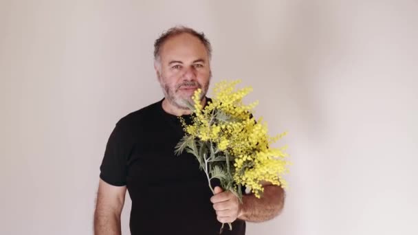 Handsom homme barbu mature souriant et donnant fleur de mimosa au spectateur. Homme avec bouquet.Concept de vacances. Mouvement lent. - Séquence, vidéo