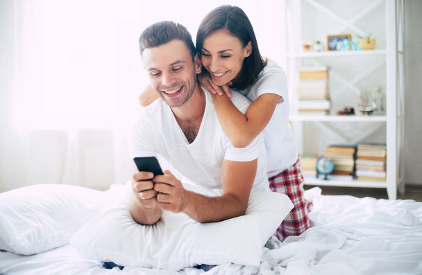 Excited and happy young smiling beautiful couple in love relaxing at home on the bed while they hug each other and having fun - Zdjęcie, obraz