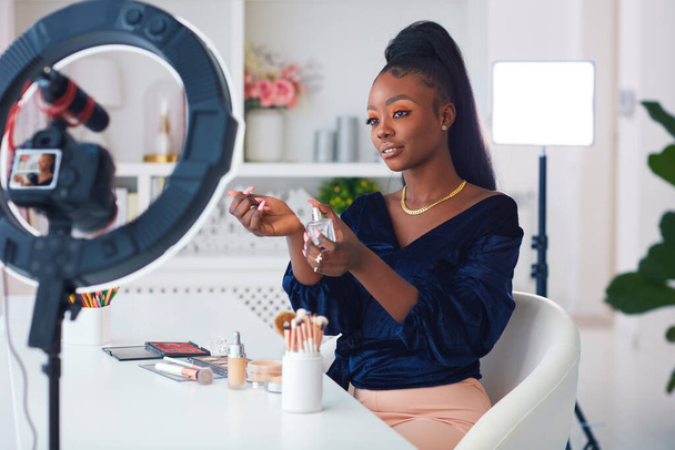 happy young african american woman streaming a beauty vlog from home, online content creator applying a perfume fragrance - Photo, Image