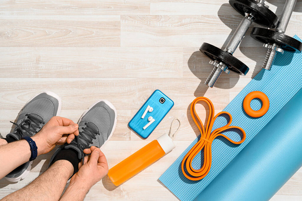 Workout at home concept. Overhead view of a man tying sport shoes with fitness equipment and smartphone on wooden floor. Morning training, sunlight. Place for text, top view. - Photo, Image