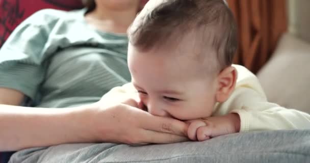 Een jonge moeder houdt een mooie zeven maanden oude zoon vast die glimlacht en knaagt aan vingers en moeders hand. jeukende tanden van een pasgeborene, tandjes krijgen - Video