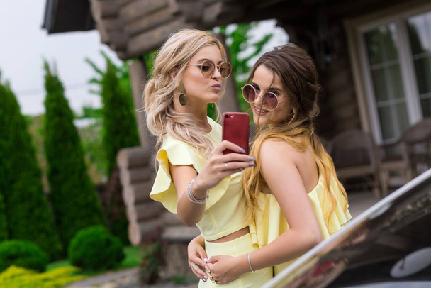 Portrait of two pretty young bridesmaids in yellow dresses near by a fashionable wooden house. People lifestyle concept. Doing selfie shot on mobile phone smiling and sending air kiss - Foto, imagen