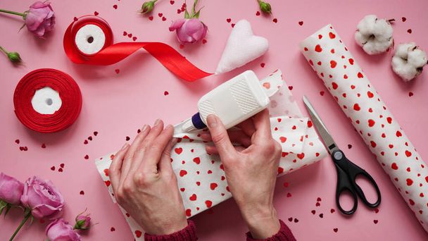 Step 4.Step-by-step instructions for wrapping gifts for Valentine's Day. Woman wraps a gift with wrapping white paper in heart and seals the edges with glue on a pink background top view, flat lay. - Photo, Image