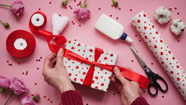 Stap 10.Stap-voor-stap instructies voor het inpakken van geschenken voor Valentijnsdag. Vrouw wikkelt een geschenk met omwikkeld wit papier in het hart en verzegelt de randen met lijm op een roze achtergrond bovenaanzicht, plat lay. - Foto, afbeelding