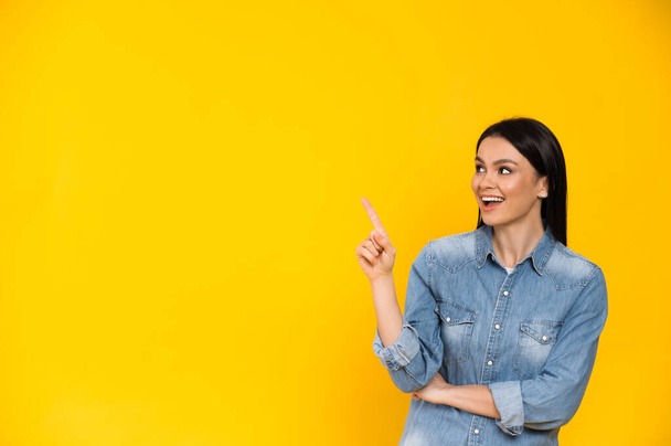 Фото збудженої досить кавказької молодої жінки, вказує пальцем на порожній простір, стоїть на ізольованому помаранчевому кольоровому фоні і посміхається
 - Фото, зображення