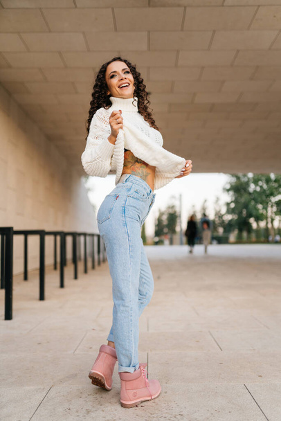 Slender beautiful brunette in blue jeans dances slightly lifting the edge of a white sweater - Фото, зображення