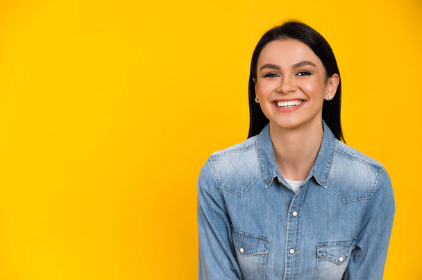 Portrait of a happy smiling caucasian young brunette woman dressed in stylish denim shirt, stands on isolated orange background and looks directly at the camera - Photo, Image