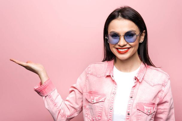 Portrait of excited caucasian pretty girl dressed in trendy clothes and sunglasses, pointing hand at blank space and smiles, stands on isolated pastel pink background. - Valokuva, kuva