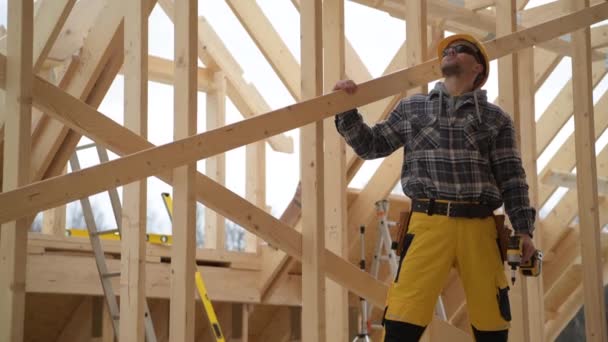Biały robotnik budowlany po czterdziestce spogląda w górę na dachu drewnianej konstrukcji szkieletowej nowo wybudowanego domu. - Materiał filmowy, wideo