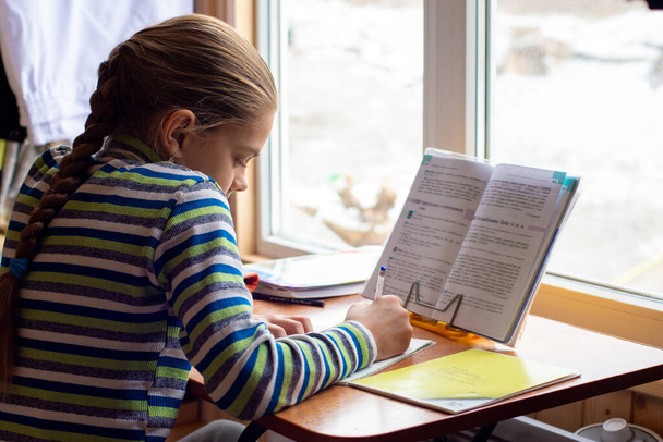 Η μαθήτρια κάθεται στο τραπέζι δίπλα στο παράθυρο και διαβάζει. - Φωτογραφία, εικόνα