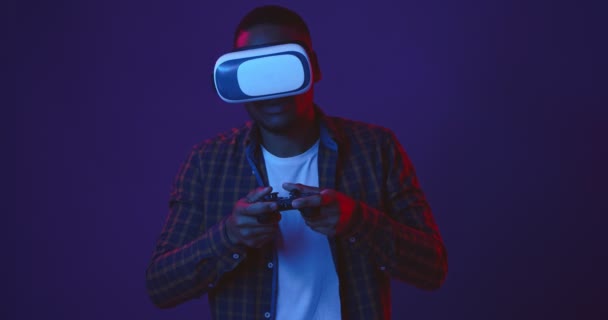 Τζόγος. Νεαρός μαύρος που παίζει βιντεοπαιχνίδια με χειριστήριο και ακουστικά εικονικής πραγματικότητας, φόντο νέον φώτα - Πλάνα, βίντεο
