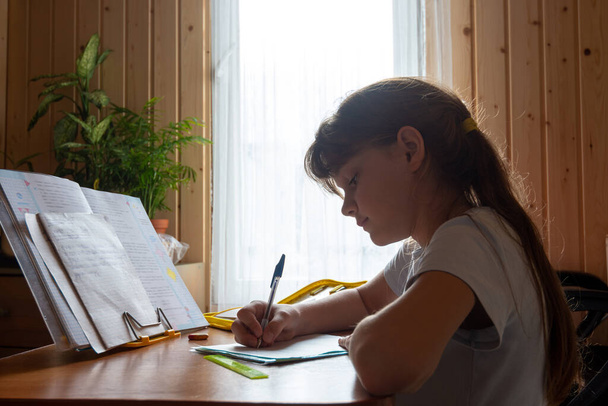 Девушка делает домашнюю работу, сидя за столом в комнате загородного дома - Фото, изображение