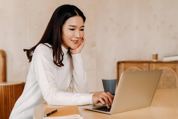Young teenage Asian woman wears sweater with pretty smile in living room. Happy and successful girl using laptop with notebook.Online technology concept. Winter portrait fashion lifestyle. Copy space - Фото, изображение