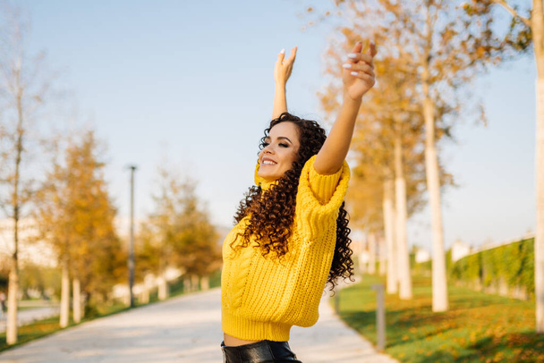 In the alley of a desert park, a girl in a bright yellow sweater spread her arms to the sides and up enjoys a sunny day and smiles at the sun - Foto, immagini