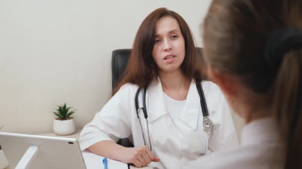 Family doctor is talking to patient in office. Doctor and patient shake hands. Smart female doctor in white coat and stethoscope consulting patient in hospital office. Therapist works with a sick girl - Footage, Video