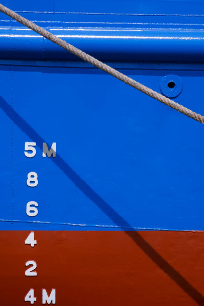 垂直フレーム内の古い巨大なファイバーロープの背後にある青と赤の防錆船船体の側面にマーク番号をドラフト - 写真・画像