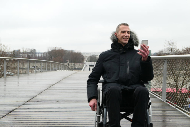 Έννοια των ατόμων με αναπηρία. Ένας άντρας σε αναπηρικό καροτσάκι έξω στο δρόμο. Άτομα που χρησιμοποιούν τεχνολογία με smartphone. - Φωτογραφία, εικόνα