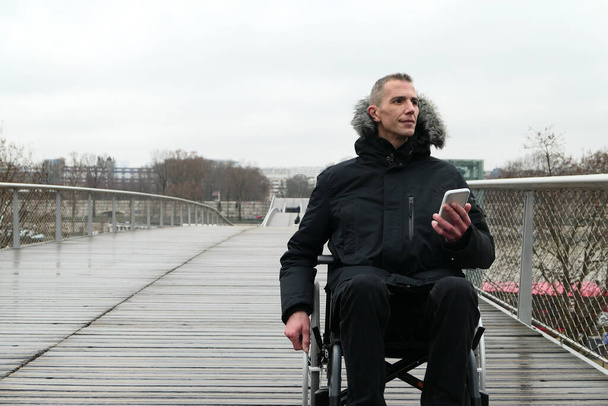 Έννοια των ατόμων με αναπηρία. Ένας άντρας σε αναπηρικό καροτσάκι έξω στο δρόμο. Άτομα που χρησιμοποιούν τεχνολογία με smartphone. - Φωτογραφία, εικόνα