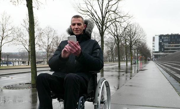 Concept of disabled person. Man in a wheelchair outside in the street. People using technology with smartphone. - Photo, Image