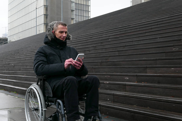 Vammaisen käsite. Mies pyörätuolissa kadun ulkopuolella portaiden edessä. Ihmiset käyttävät teknologiaa älypuhelimella. - Valokuva, kuva