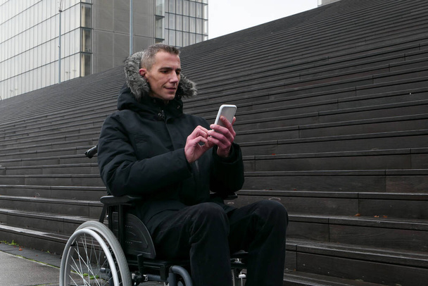 Pojęcie osoby niepełnosprawnej. Mężczyzna na wózku inwalidzkim na ulicy przed schodami. Ludzie używający technologii ze smartfonem. - Zdjęcie, obraz