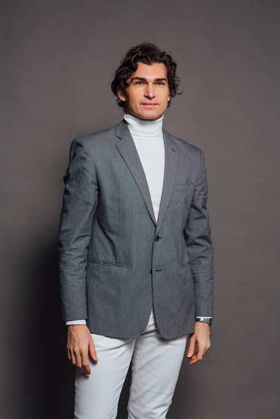 Fashion portrait of a tall handsome man dressed in white turtleneck, white jeans and grey jacket posing on the grey background - Foto, Bild