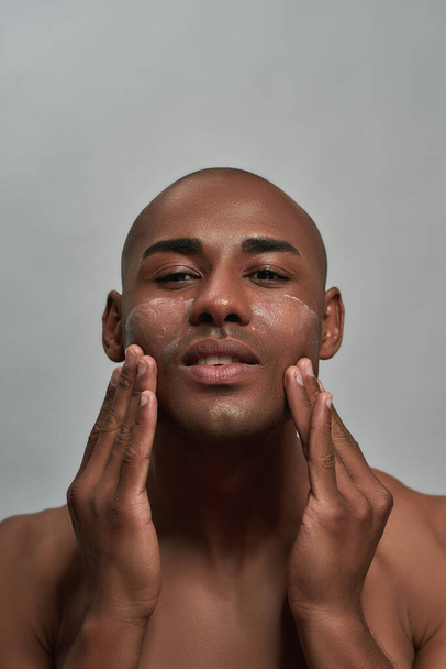 Portrait of handsome young african american man looking at camera, applying cream on his face, posing isolated over gray background - Zdjęcie, obraz