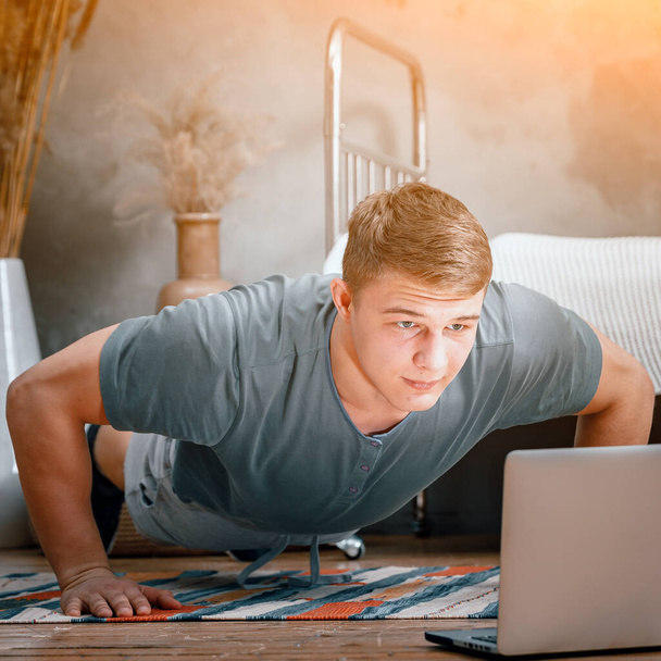 The young man goes in for sports at home. The athlete does push-ups and watches a movie and studies from a laptop in the bedroom, in the background a bed, a vase, a carpet - Photo, Image