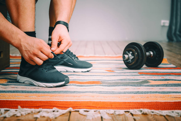 Primo piano di un uomo che lega i lacci delle scarpe da ginnastica sportive nere, un orologio nero sportivo sulla mano e manubri di metallo nelle vicinanze - Foto, immagini