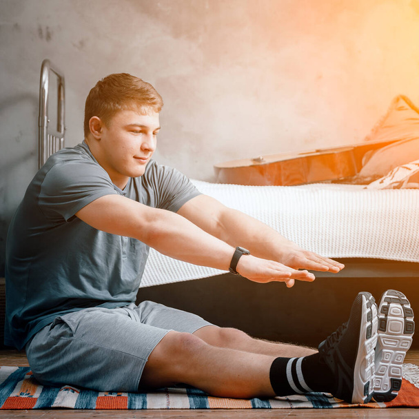 Een jongeman gaat thuis sporten, online trainen via de telefoon. De atleet strekt zich uit in de slaapkamer, op de achtergrond is er een bed, een tapijt. - Foto, afbeelding