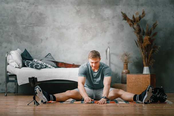 Сильный мужчина занимается спортом дома. Веселый спортивный человек с светлыми волосами тянется к ноге - Фото, изображение