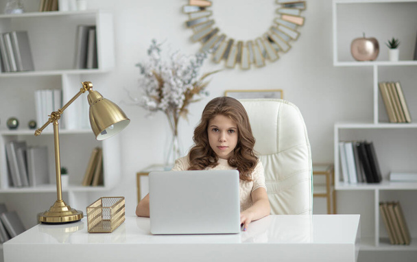 Beautiful teenage girl with blond long curly hair sits at a laptop learns online during a pandemic in light home interior. Distance learning concept. - Foto, imagen