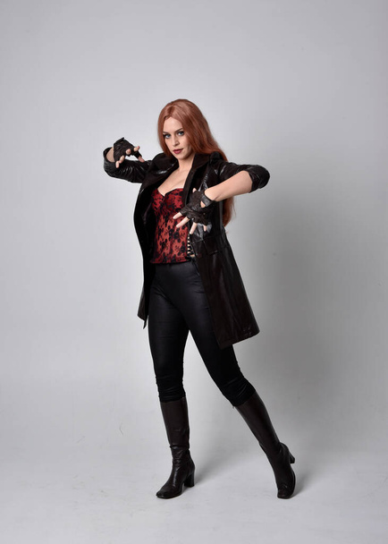 Ganzkörperporträt einer Frau mit langen roten Haaren, die dunklen Ledermantel, Korsett und Stiefel trägt. Stehende Pose vor Studiohintergrund mit witzigen Handgesten. - Foto, Bild