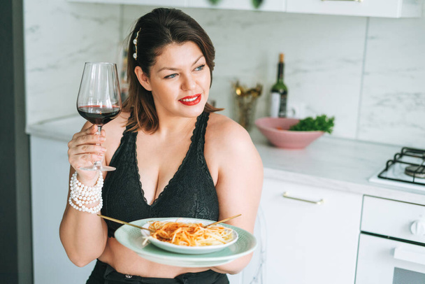 Mooie sensuele brunette jonge vrouw plus size lichaam positief met schotel pasta en glas wijn genieten van haar leven thuis - Foto, afbeelding