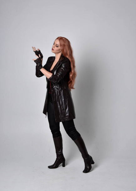 full length portrait of woman with long red hair wearing dark leather coat, corset and boots. Standing pose facing front on wit hand gestures against a  studio background. - Fotografie, Obrázek