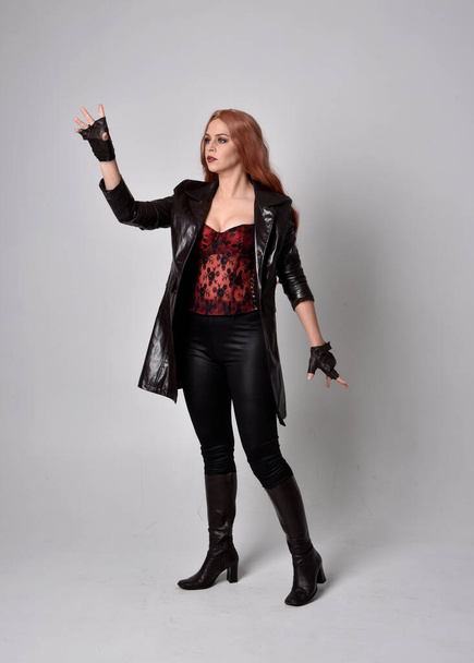 full length portrait of woman with long red hair wearing dark leather coat, corset and boots. Standing pose facing front on wit hand gestures against a  studio background. - Photo, Image