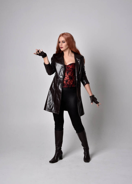 full length portrait of woman with long red hair wearing dark leather coat, corset and boots. Standing pose facing front on wit hand gestures against a  studio background. - Photo, Image
