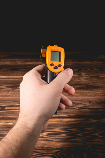 Un hombre sostiene un pirómetro amarillo-negro sobre un fondo de madera. Dispositivo para la medición de temperatura sin contacto. Estudio foto. - Foto, imagen