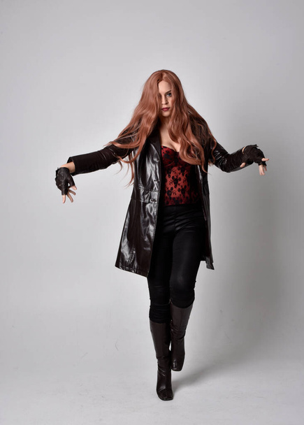Uzun kızıl saçlı, koyu deri ceketli, korseli ve çizmeli bir kadın portresi. Stüdyonun arka planında el hareketlerinin önünde durmak. - Fotoğraf, Görsel