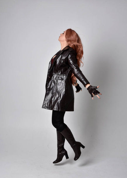 full length portrait of woman with long red hair wearing dark leather coat, corset and boots. Standing pose facing front on wit hand gestures against a  studio background. - Φωτογραφία, εικόνα