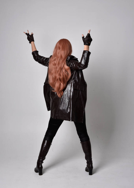 full length portrait of girl with long red hair wearing dark leather coat, corset and boots. Standing pose facing back view with  magical hand gestures against a  studio background. - Fotoğraf, Görsel