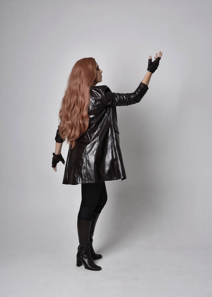 portrét dívky s dlouhými zrzavými vlasy v tmavém koženém kabátě, korzetu a botách. Stojící póza obrácený zpět pohled s kouzelnými gesty rukou na pozadí studia. - Fotografie, Obrázek