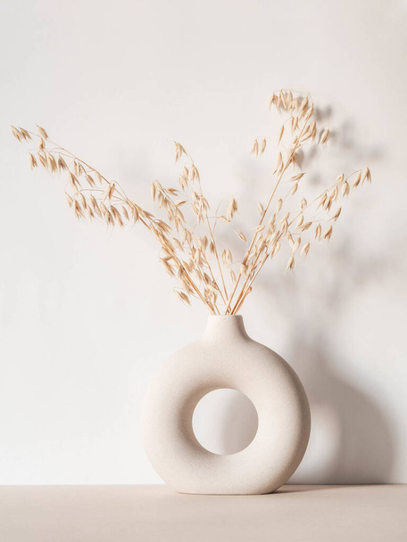 Round stylish ceramic vase with dried oats plant casting shadows on the wall. Front view. Copy space - 写真・画像