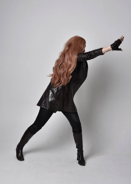 portrait complet de fille aux longs cheveux roux portant un manteau en cuir foncé, un corset et des bottes. Pose debout face à l'arrière avec des gestes de main magiques sur fond de studio. - Photo, image