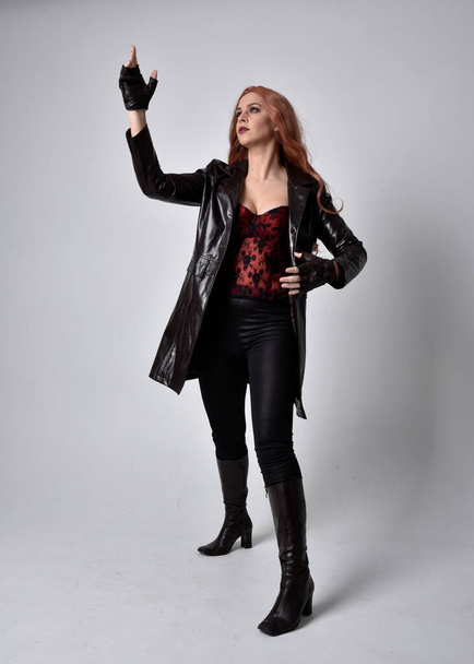 full length portrait of girl with long red hair wearing dark leather coat, corset and boots. Standing pose facing front on with  magical hand gestures against a  studio background, low camera angle. - Photo, Image