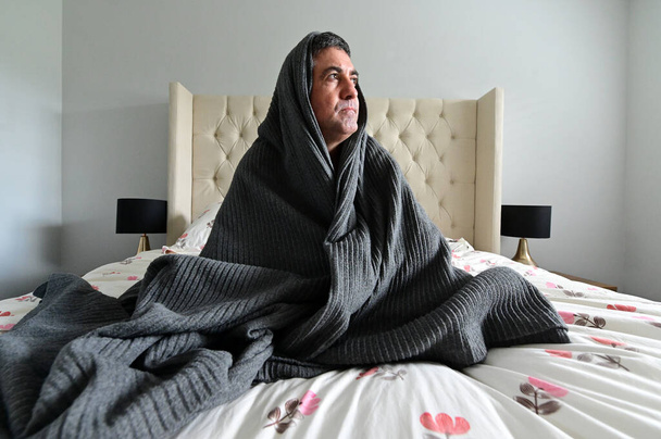 Bang volwassen man (leeftijd 40-50) bedekt met deken zitten op bed in de slaapkamer. Mentale ziekte concept.Echte mensen. Kopieerruimte - Foto, afbeelding