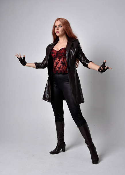 full length portrait of girl with long red hair wearing dark leather coat, corset and boots. Standing pose facing front on with  magical hand gestures against a  studio background, low camera angle. - Foto, afbeelding