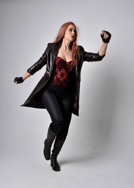 full length portrait of girl with long red hair wearing dark leather coat, corset and boots. Standing pose facing front on with  magical hand gestures against a  studio background, low camera angle. - Valokuva, kuva
