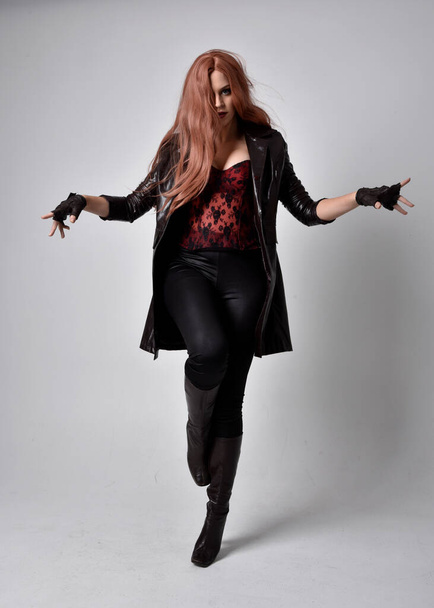 full length portrait of girl with long red hair wearing dark leather coat, corset and boots. Standing pose facing front on with  magical hand gestures against a  studio background, low camera angle. - Foto, Imagen