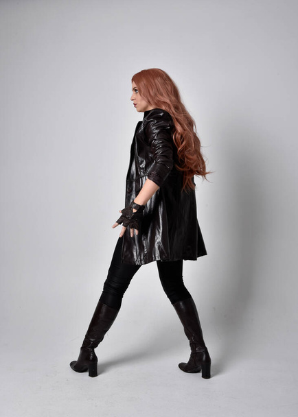 full length portrait of girl with long red hair wearing dark leather coat, corset and boots. Standing pose facing front on with  magical hand gestures against a  studio background, low camera angle. - Photo, Image